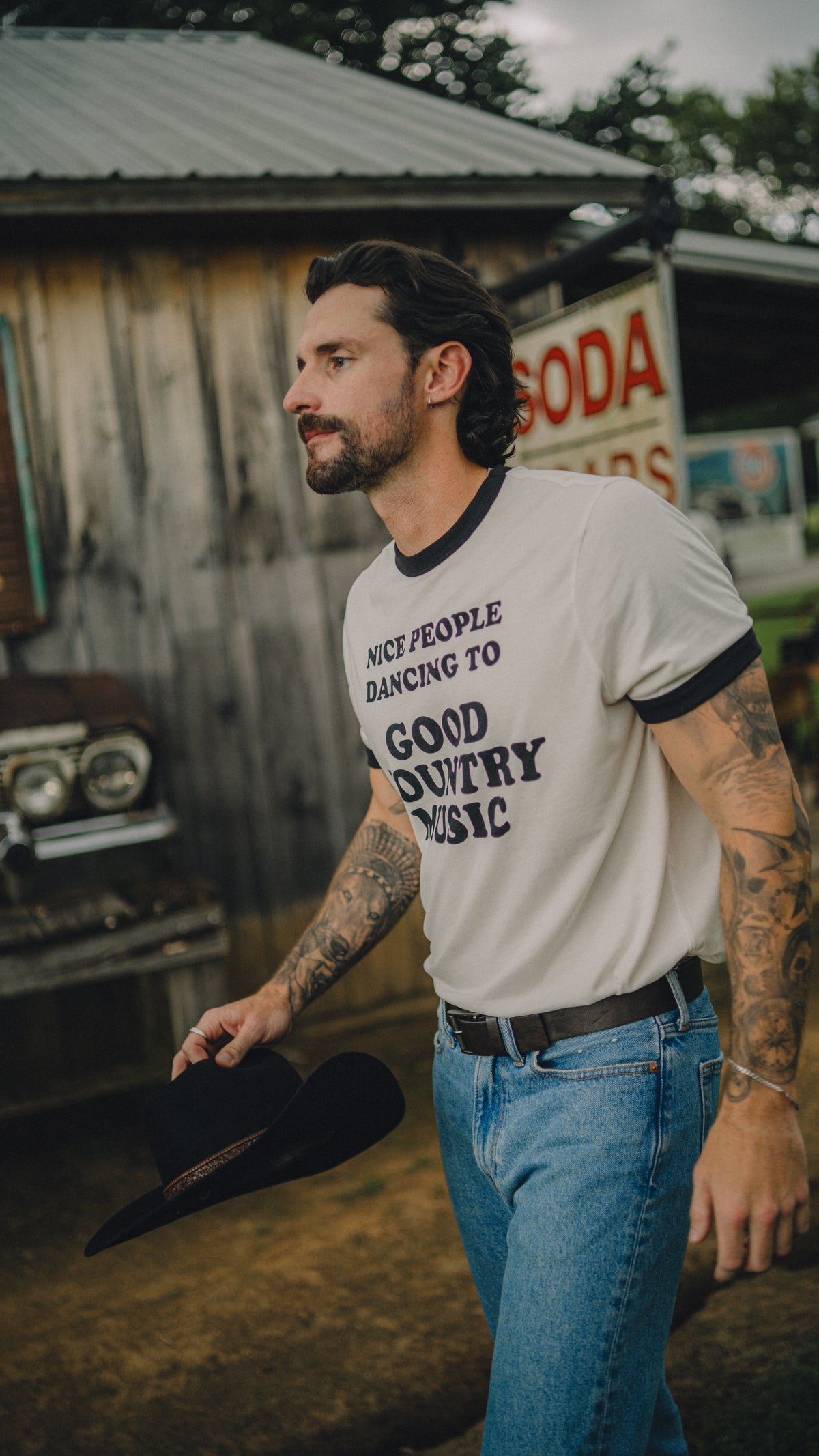 Man standing outside a rustic barn wearing the cream H Bar C x Rustler Nice People ringer T-shirt.