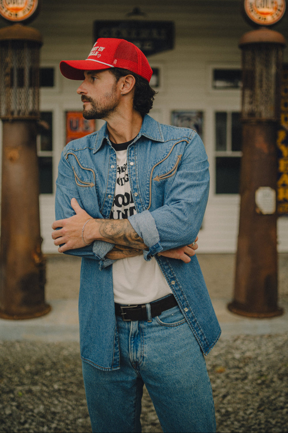 Man in a red cap wearing the Rustler x H Bar C men's denim western shirt open over a graphic tee at a vintage gas station.