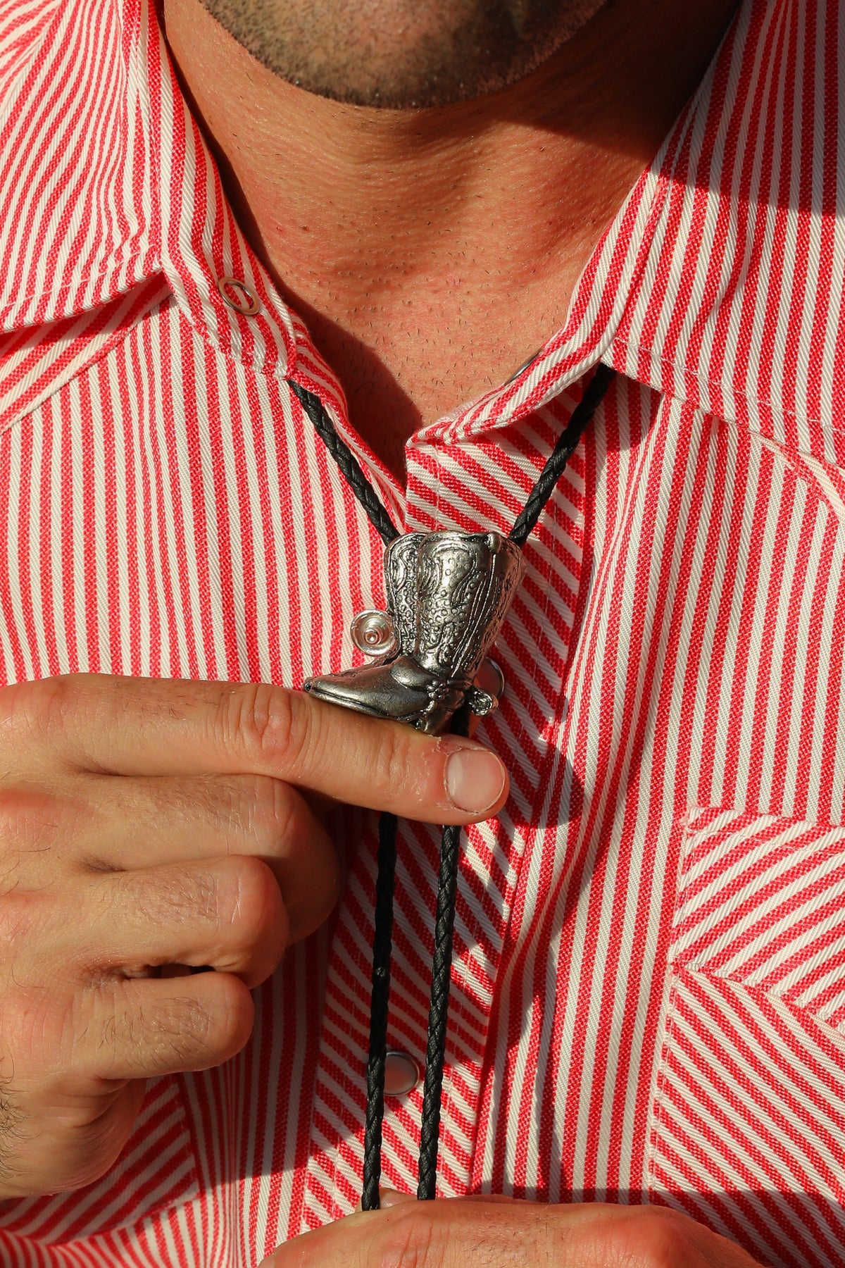 Man adjusting the H Bar C silver cowboy boot bolo tie on a red striped western shirt.
