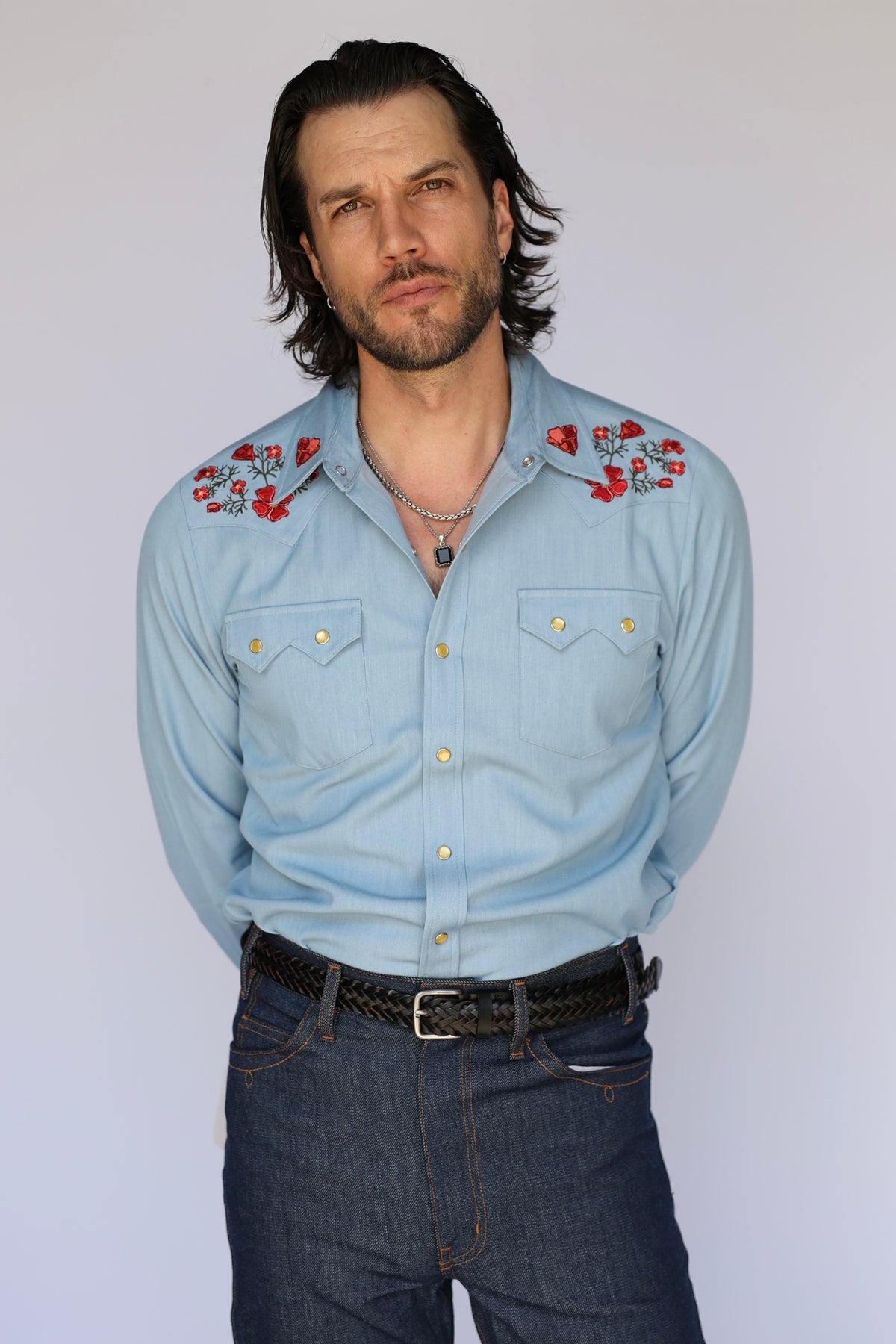 Model in the HBarC x Gibson Dove light blue chambray western shirt with red floral embroidery, worn with dark denim and a black belt.