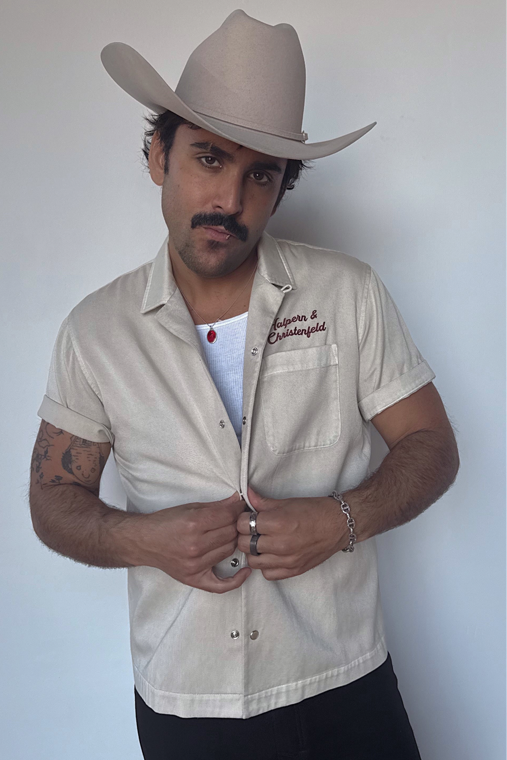 Man in a cowboy hat wearing the H Bar C Halpern & Christenfeld bowling shirt in cream with red script logo, honoring the brand's original name.