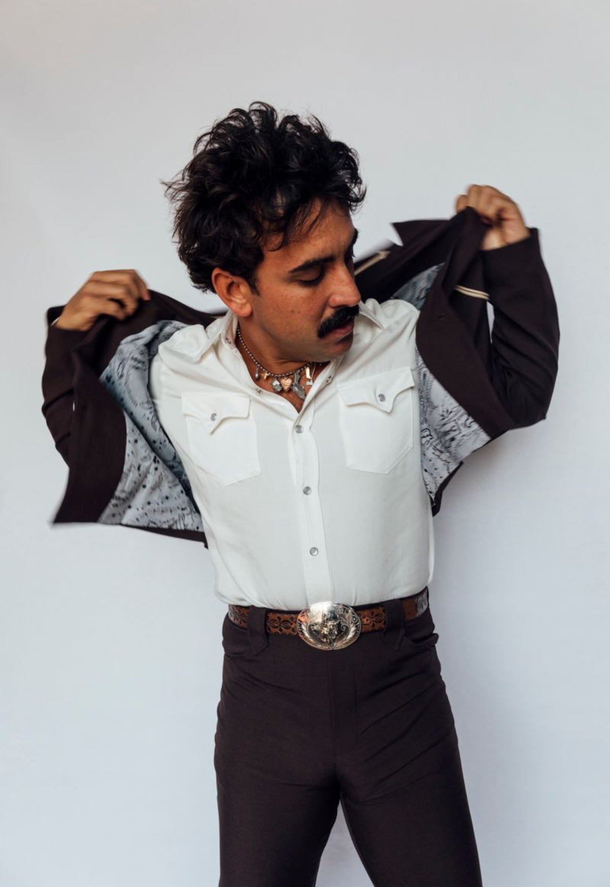 Man opening the H Bar C Calamity chocolate jacket to show the lining, worn with matching pants and silver buckle.