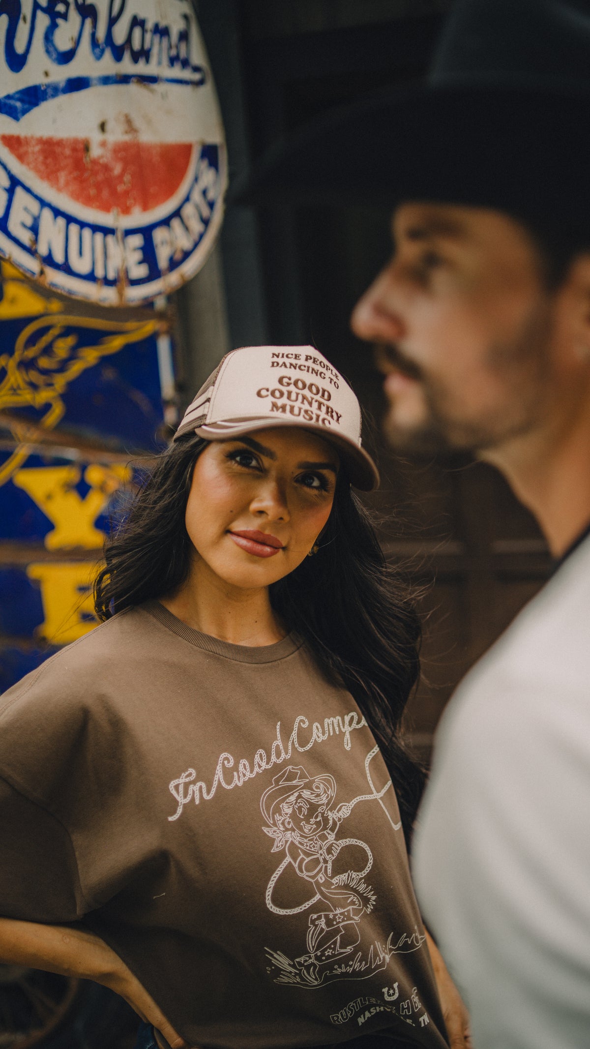 Woman wearing the H Bar C x Rustler Nice People Dancing to Good Country Music trucker cap beside a cowboy.