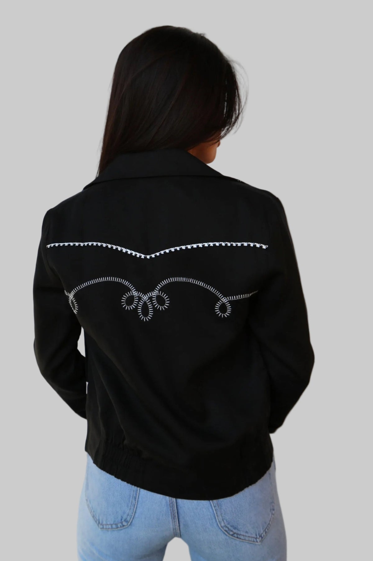 Back view of the H Bar C Bolero women's jacket in black with white chain stitch embroidery across the back yoke, worn with jeans.