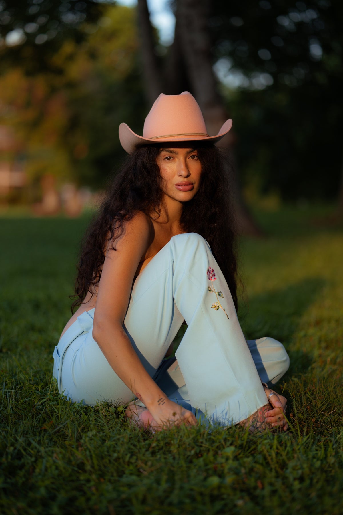 Woman seated on grass in a pink cowboy hat wearing the H Bar C Abilene pants in light blue with rose embroidery, a 1970s H Bar C replica.