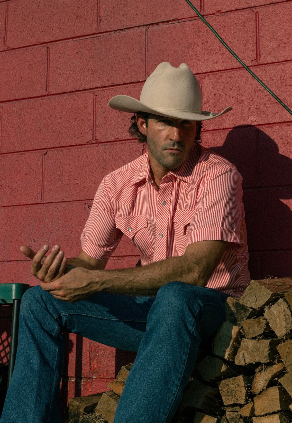 Man in a straw cowboy hat seated outside a red barn wearing the H Bar C Newport red striped short sleeve shirt.