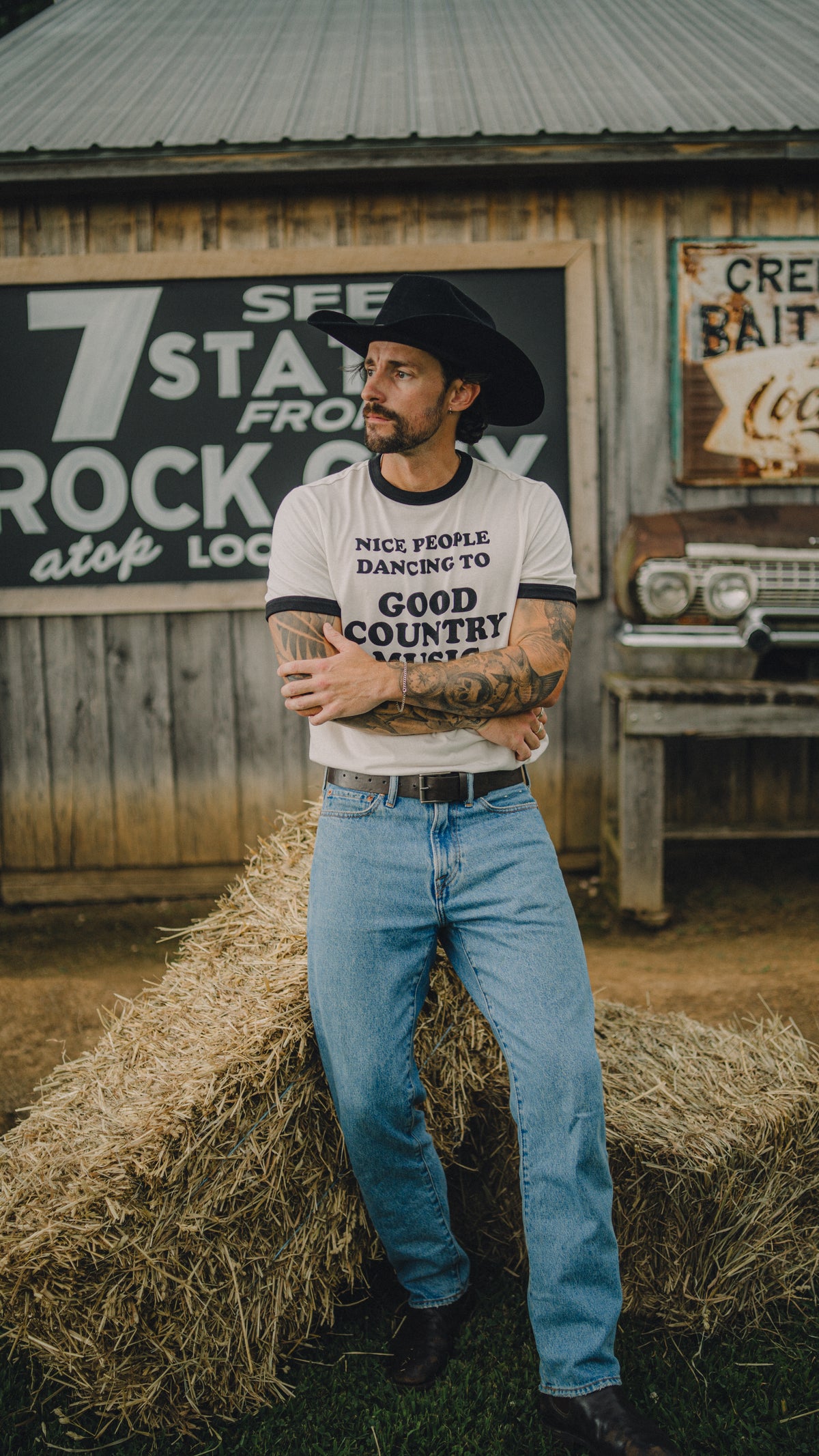 Man sitting on a hay bale outside a vintage store in the H Bar C x Rustler Nice People ringer and jeans.