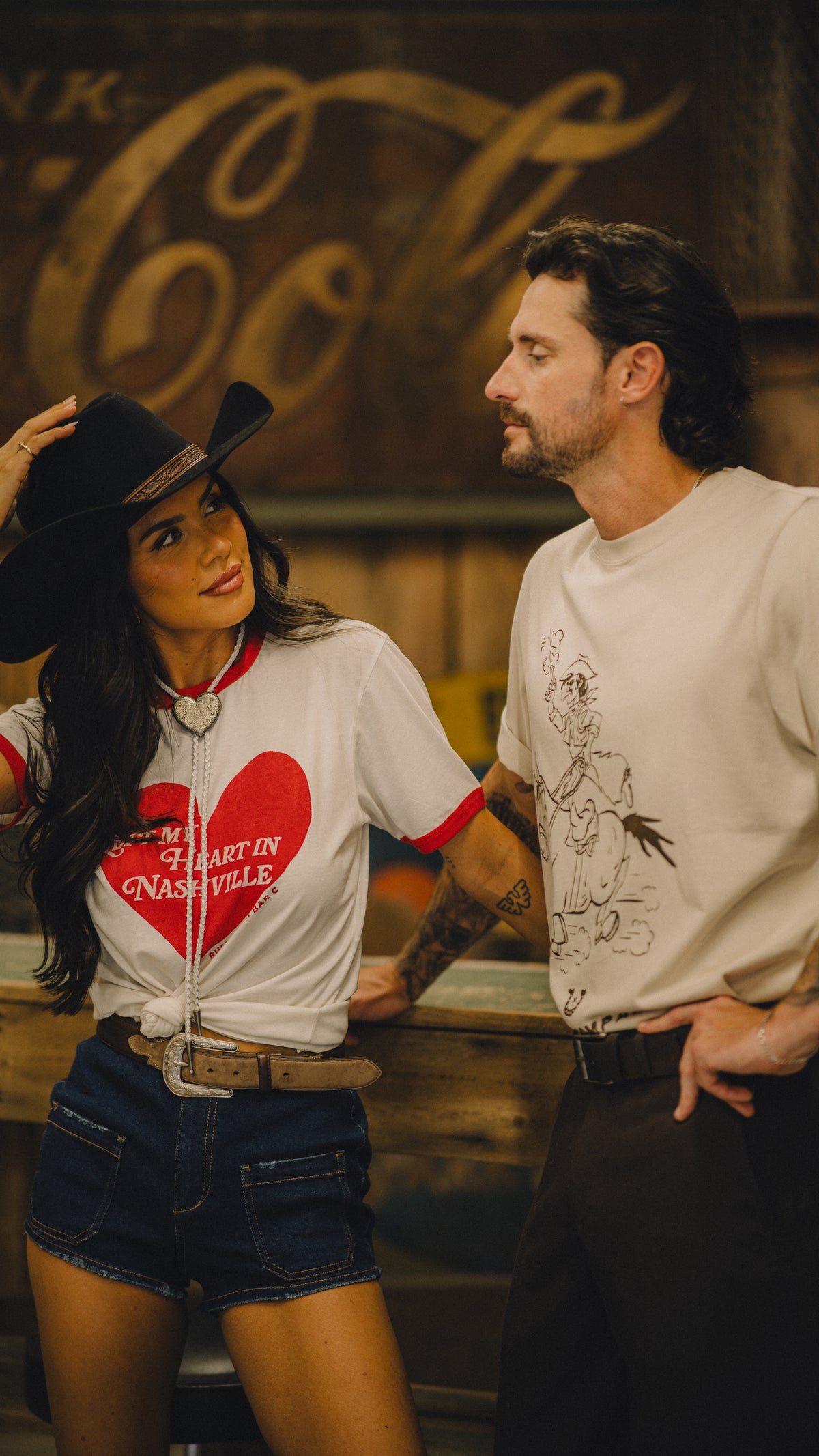 Woman in the H Bar C x Rustler Left My Heart ringer tee beside a cowboy near a vintage Coca-Cola sign.