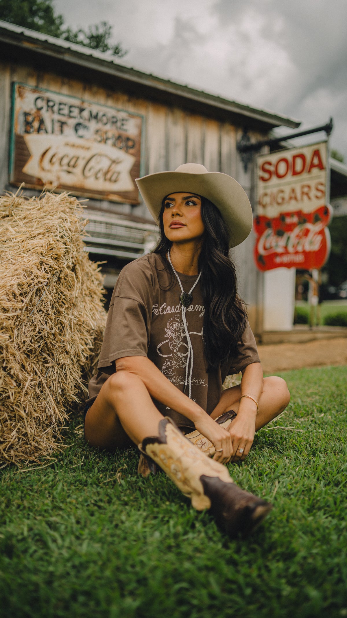 Woman seated on grass wearing the H Bar C x Rustler In Good Company women's crop tee and cowboy hat.
