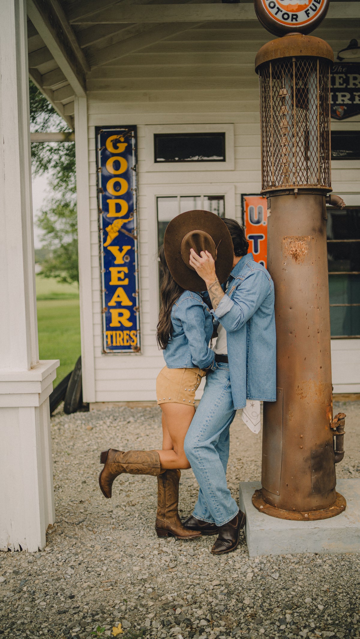 Couple in cowboy hats embracing on a vintage gas station porch, both wearing Rustler x H Bar C denim western shirts.