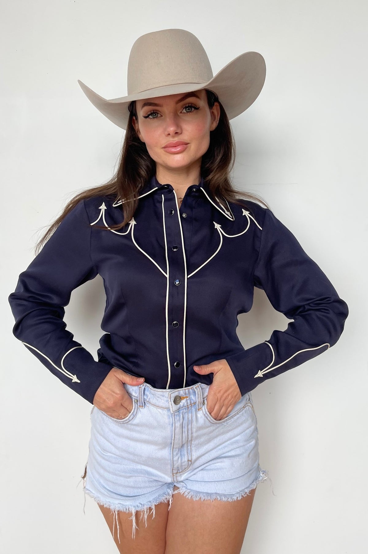 Woman in a cream cowboy hat wearing the H Bar C Lake Tahoe women's shirt in navy with white piping, tucked into denim shorts.