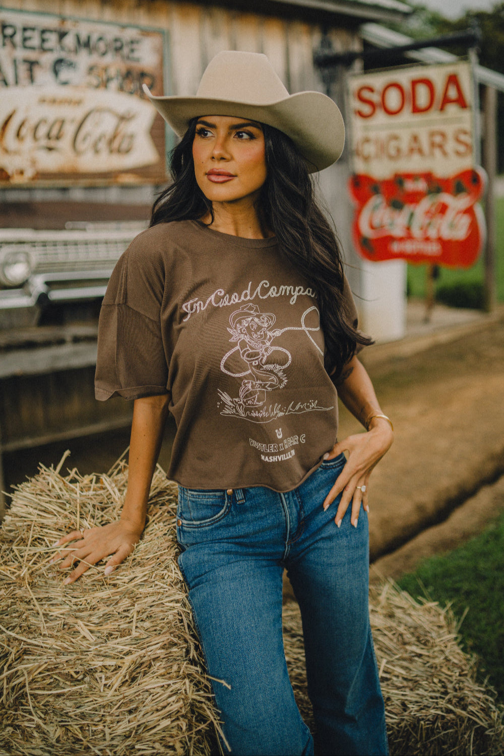Woman in a cowboy hat sitting on a hay bale outside a vintage general store wearing the H Bar C x Rustler In Good Company women's crop tee.