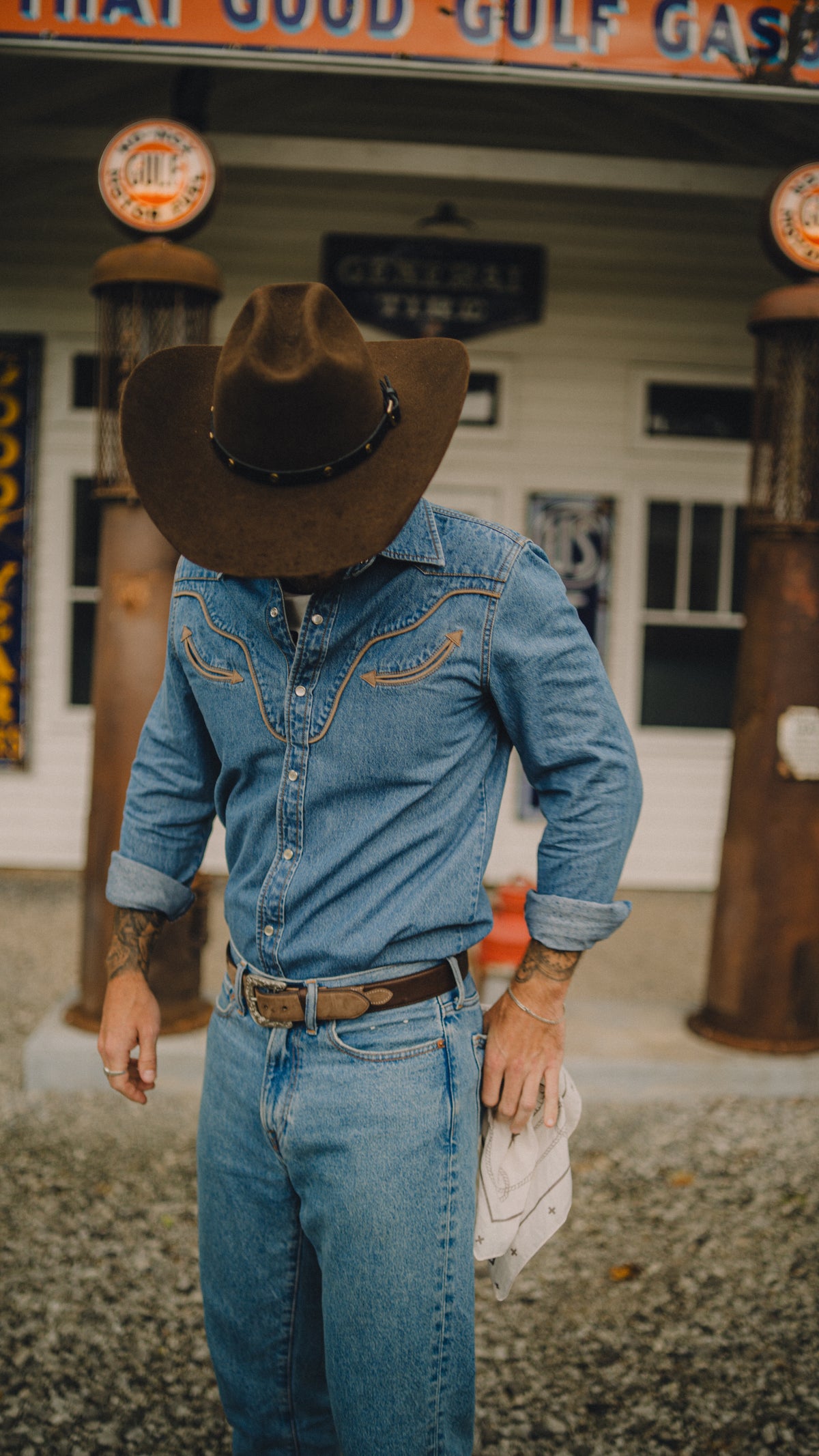 Man in a cowboy hat wearing the Rustler x H Bar C denim western shirt with jeans in front of vintage gas pumps.
