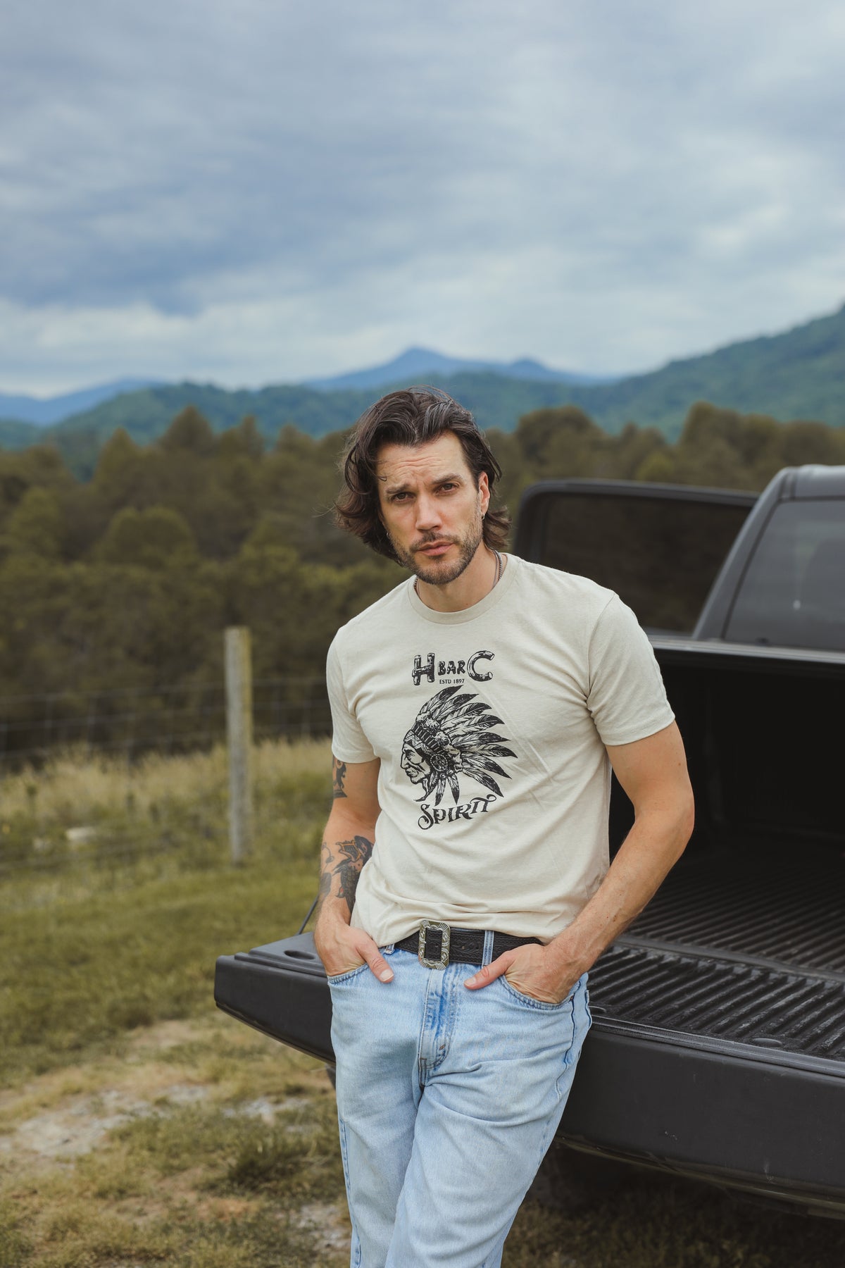Man leaning on a pickup truck in the mountains wearing the H Bar C Sand Spirit graphic tee and jeans.