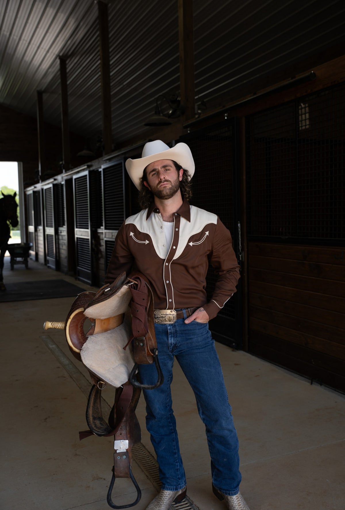 Full length view of a man holding a western saddle, wearing the H Bar C San Juan chocolate shirt and jeans in a horse barn.