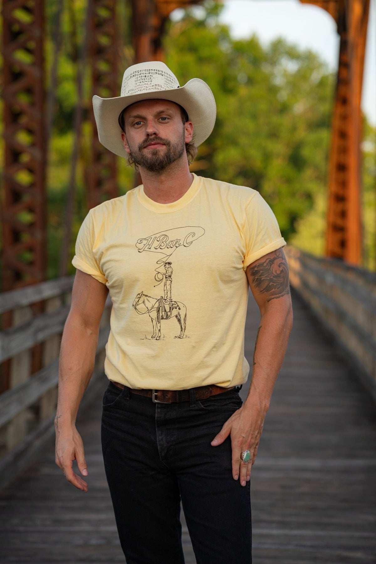 Man in a cowboy hat on an iron bridge wearing the H Bar C Tall Saddle tee in yellow, a Kate Snake Art collaboration.