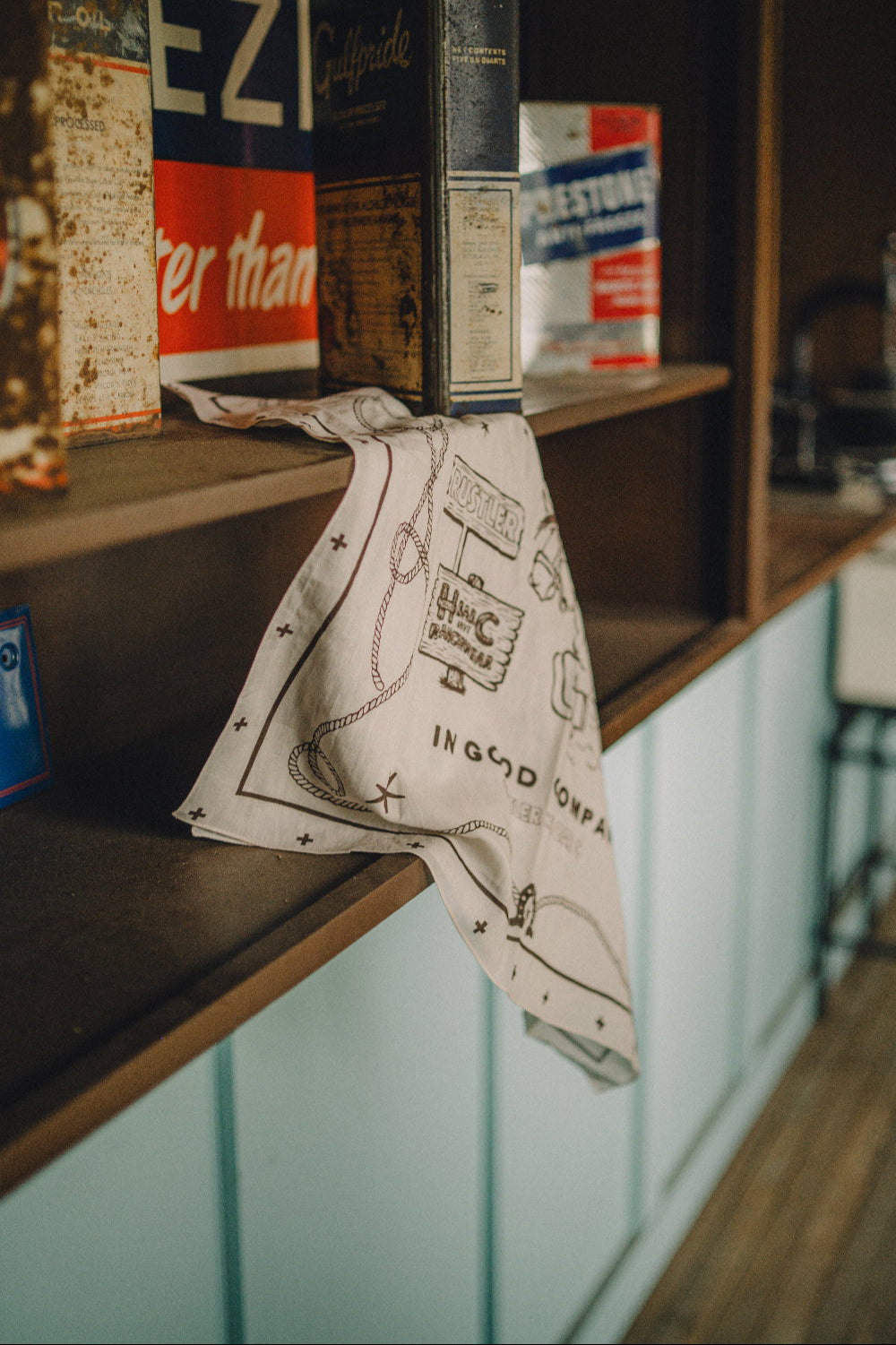 H Bar C x Rustler In Good Company bandana in cream with brown western print, hanging on a vintage store counter.