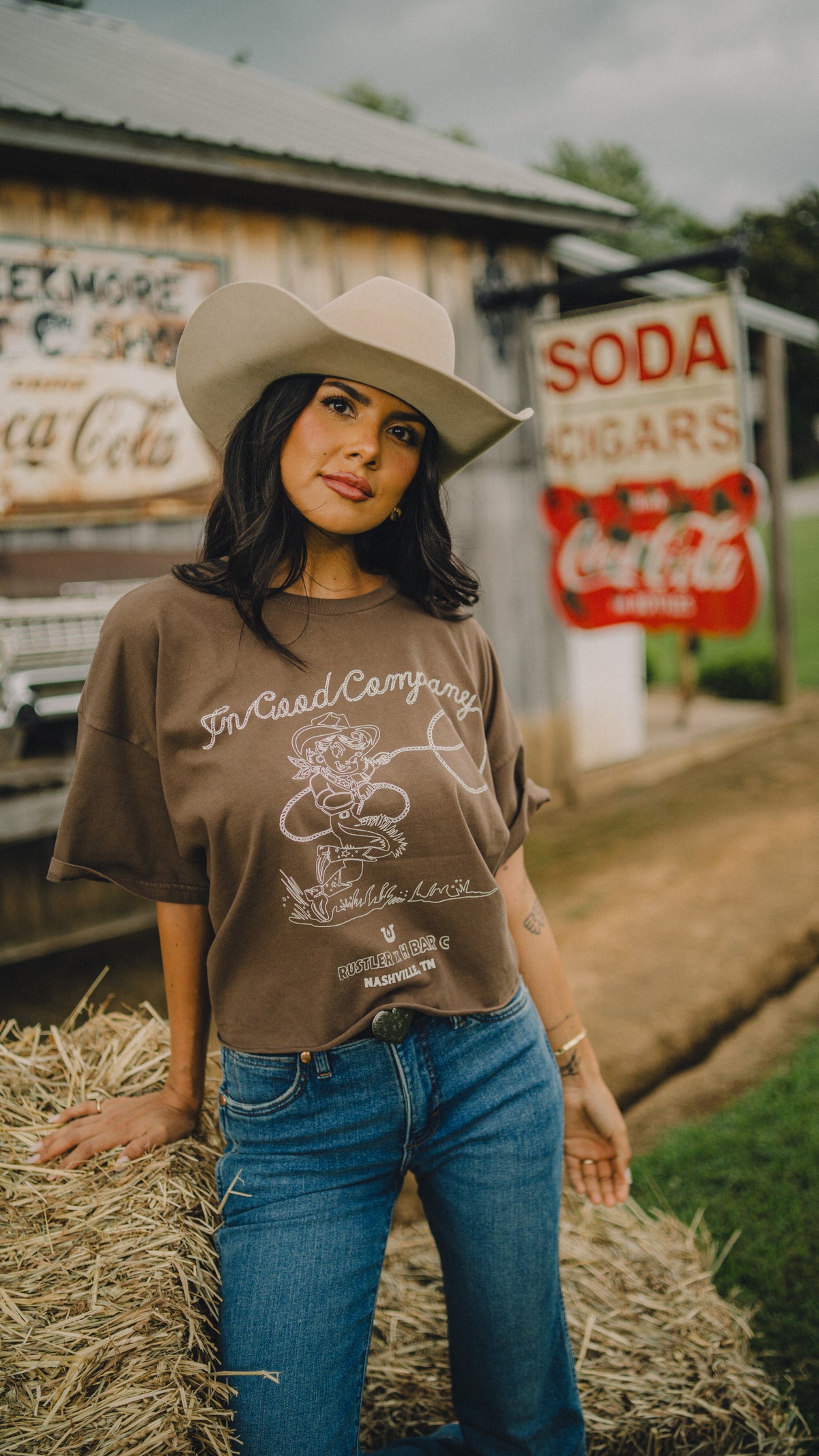 Woman wearing the brown H Bar C x Rustler In Good Company cowgirl crop tee with jeans by vintage soda signs.