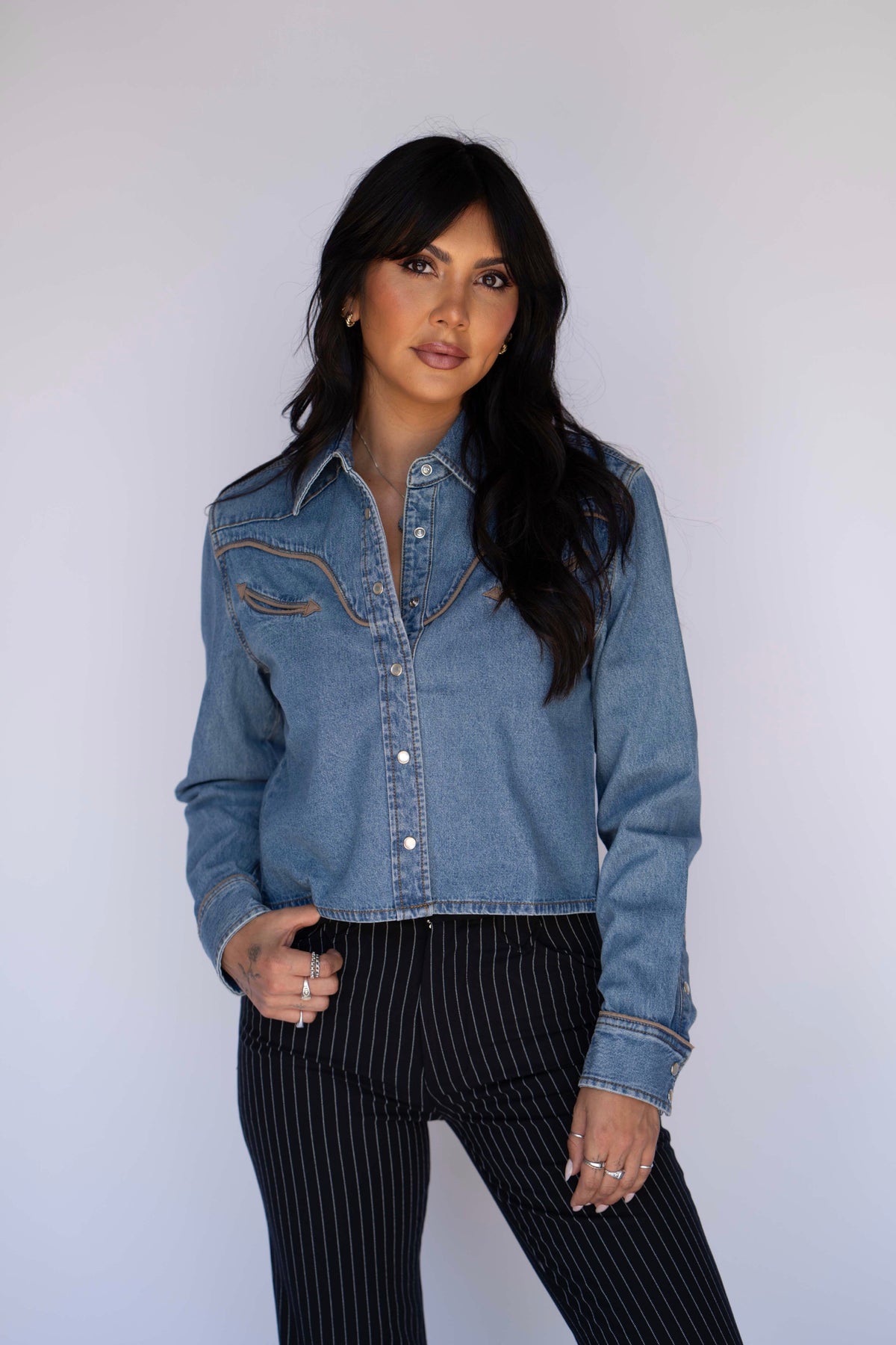 Woman wearing the Rustler x H Bar C denim cropped shirt in medium wash with tan piping and arrow pockets, styled with black pinstripe pants.