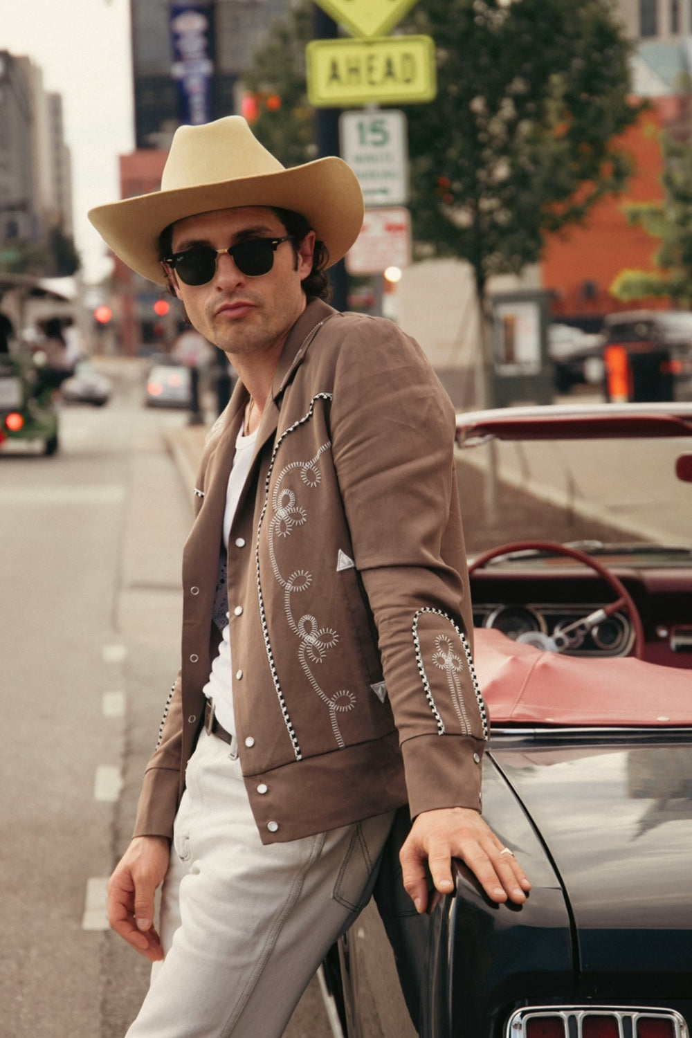 Man in a cowboy hat and sunglasses wearing the H Bar C Bolero cocoa and white jacket with white pants, leaning on a vintage car on a city street.