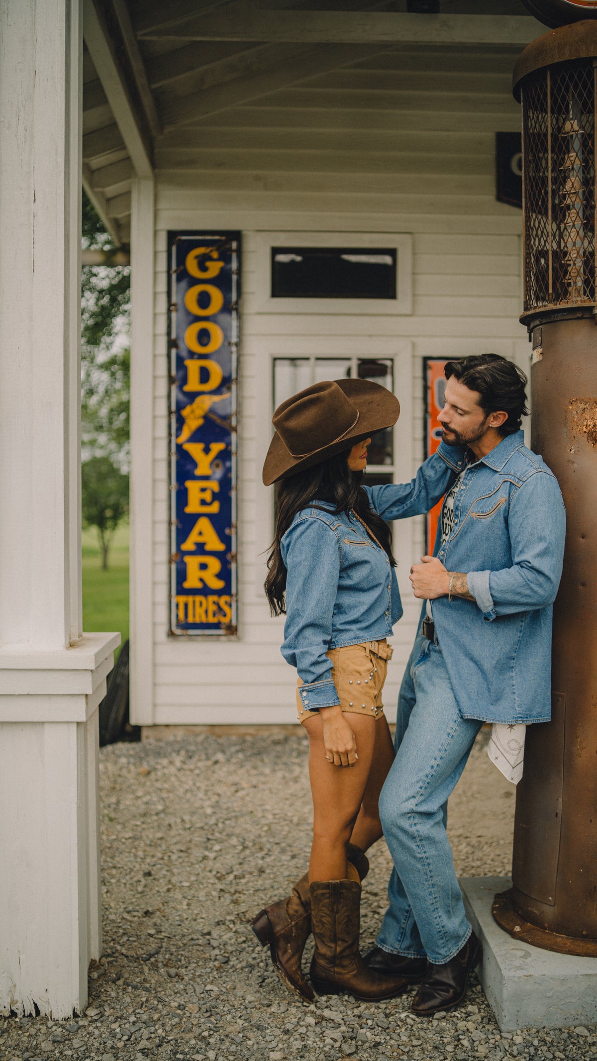 Man and woman in Rustler x H Bar C denim western shirts standing on a porch with a vintage Goodyear sign.