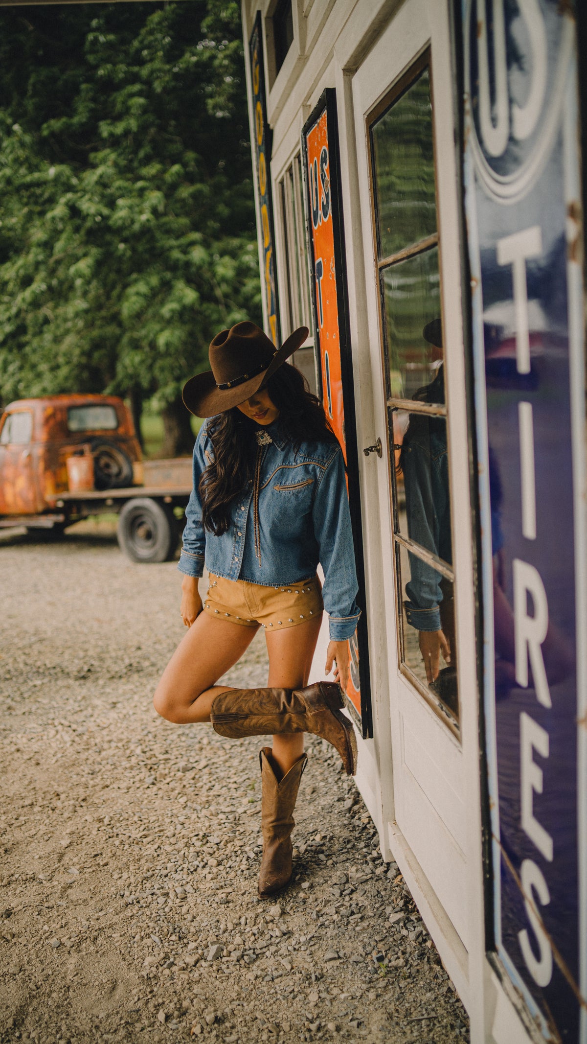 Woman in a cowboy hat and boots wearing the Rustler x H Bar C denim shirt, leaning in a vintage gas station doorway.