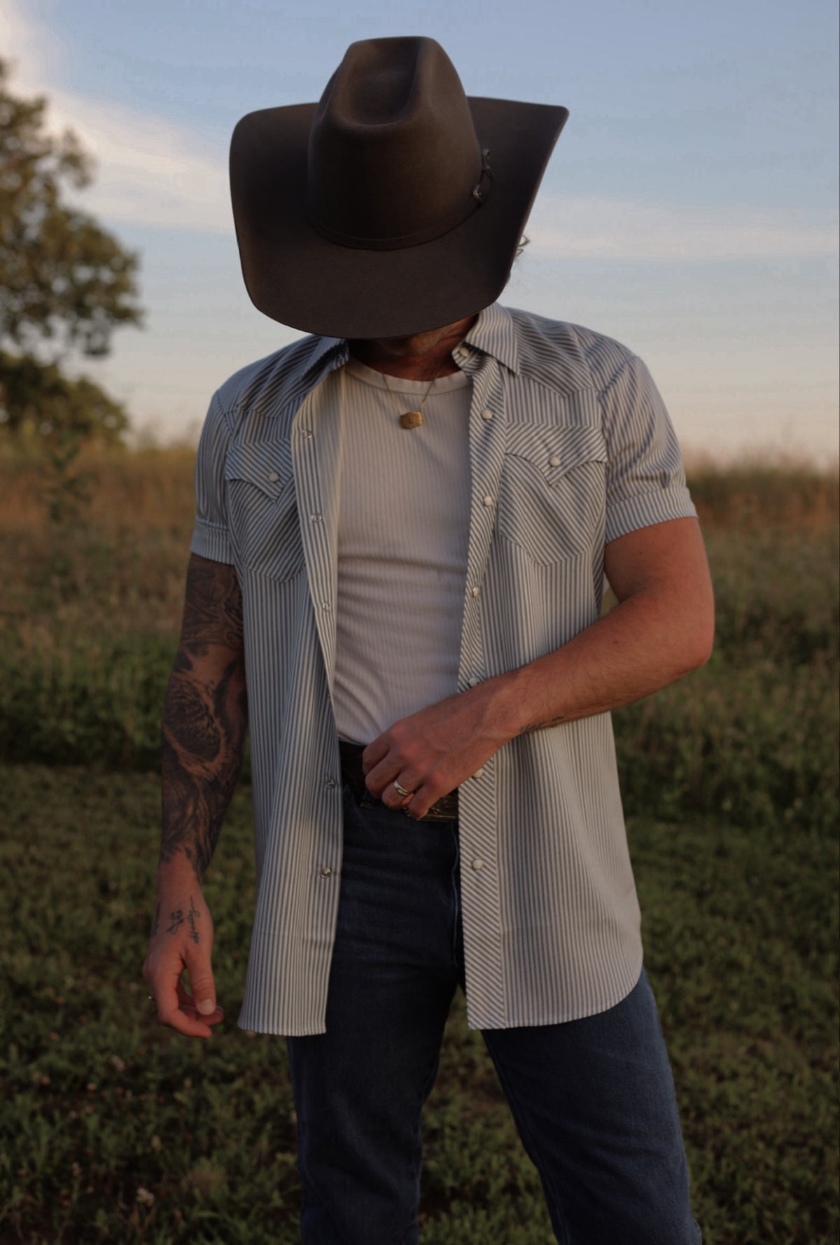 Man in a black cowboy hat wearing the H Bar C Newport gray striped short sleeve shirt open over a tee in a summer field.