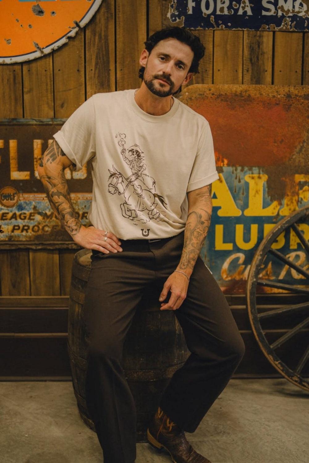 Man seated outside a vintage general store wearing the H Bar C x Rustler In Good Company men's tee in cream.