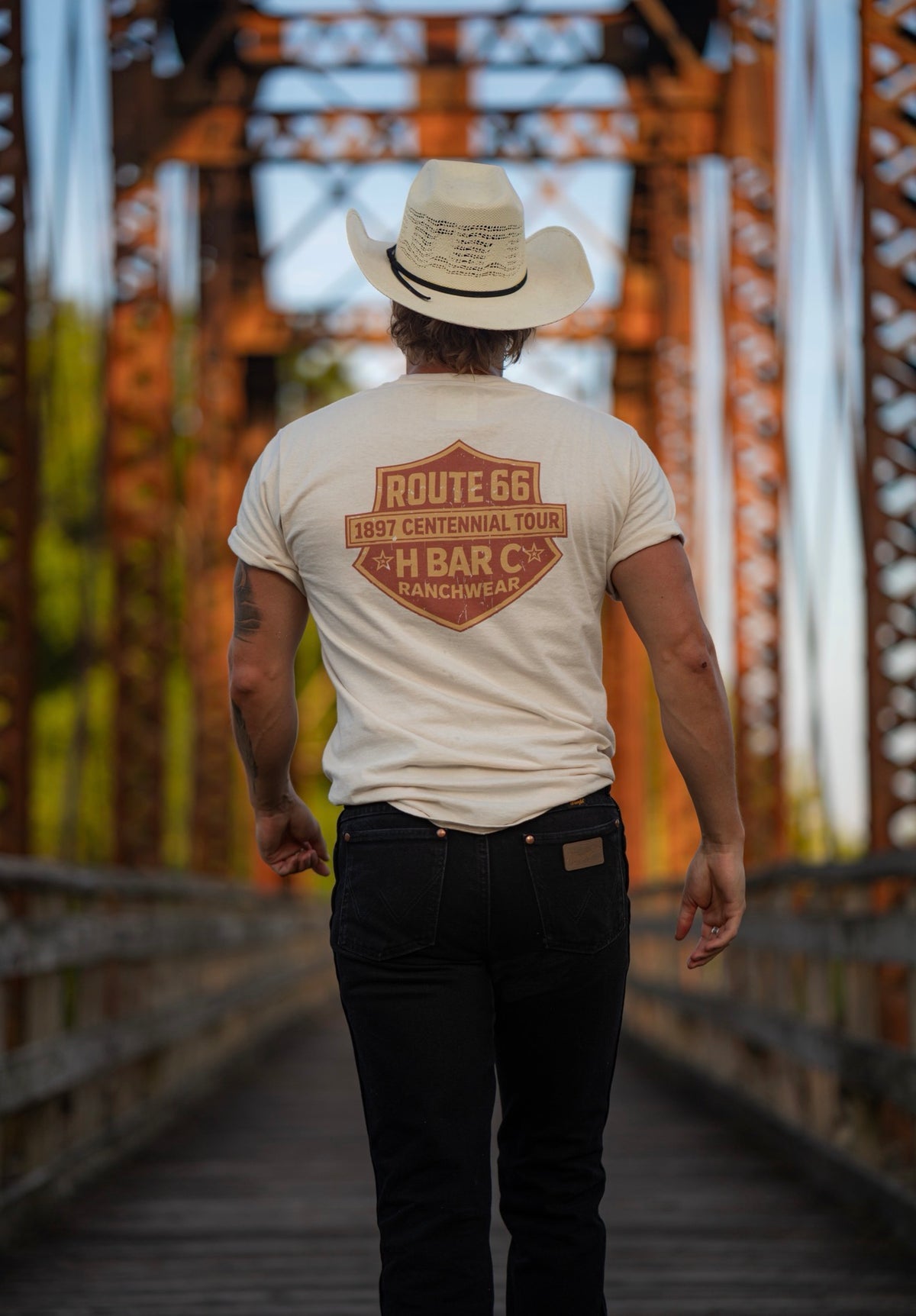 Man walking across an old truss bridge wearing the H Bar C Tulsa Shield Route 66 tee and black jeans.