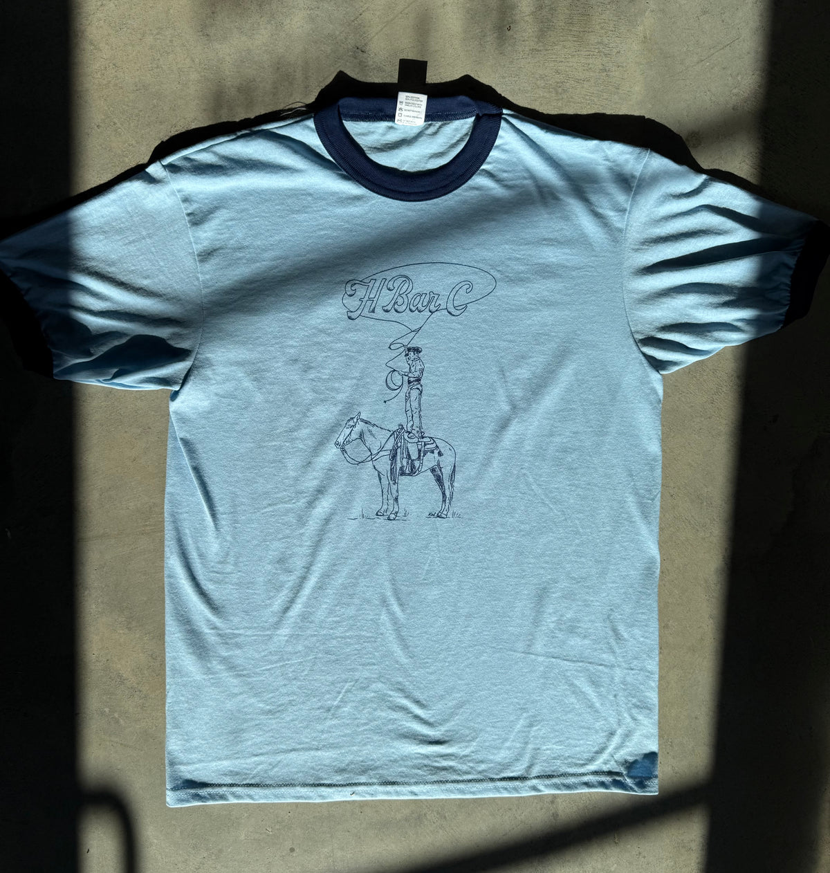 Light blue H Bar C ringer T-shirt with navy trim and Kate Snake Art trick rider graphic.