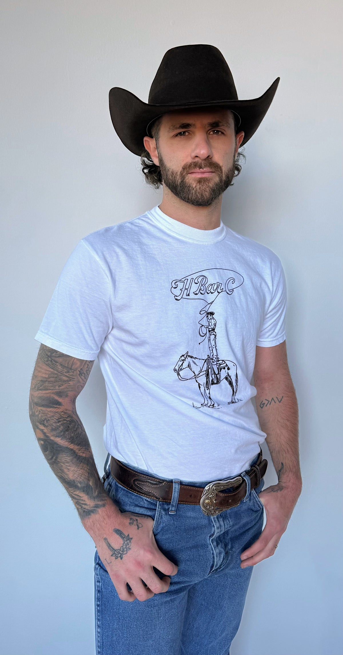 Front view of the white H Bar C Tall Saddle graphic T-shirt worn with jeans and a black hat.