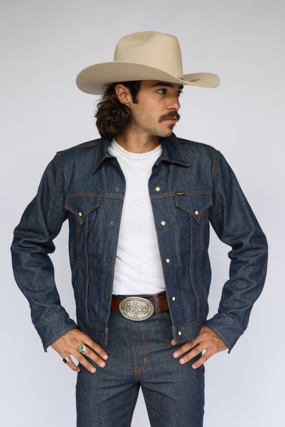 Man in a cream cowboy hat wearing the H Bar C Salina indigo trucker jacket with a white tee and silver buckle.