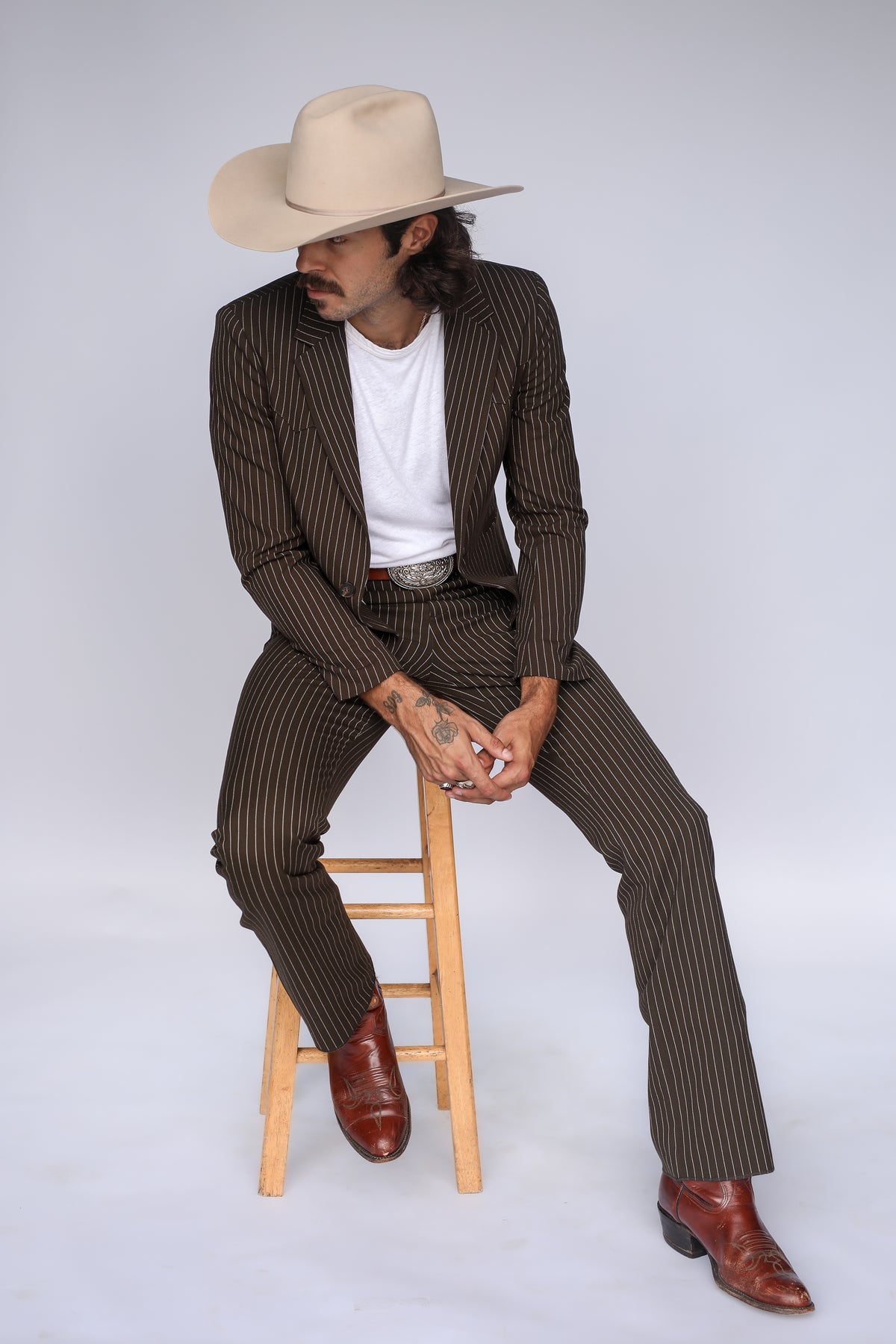 Man sitting on a wooden stool in the H Bar C Bakersfield brown pinstripe jacket and western pants.