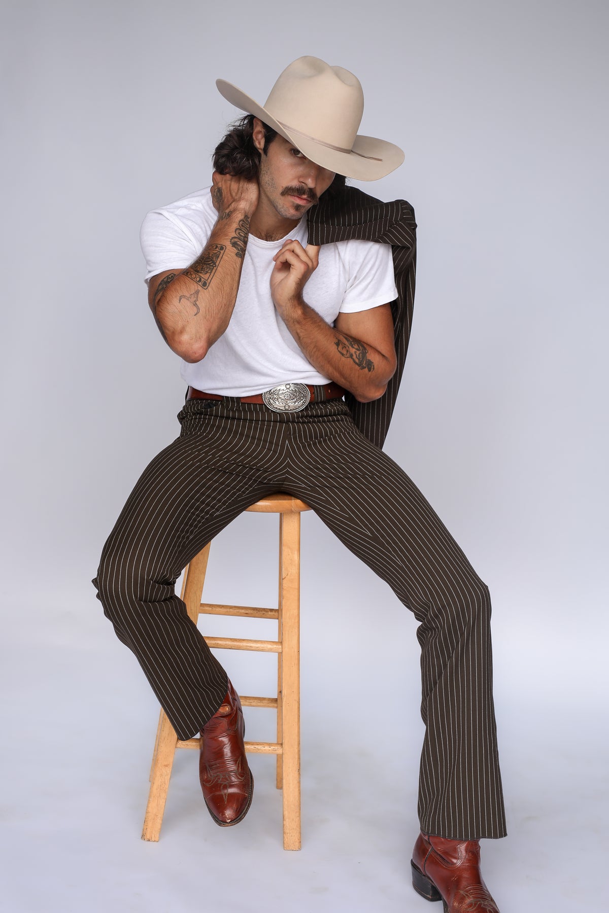 Man seated on a stool wearing the H Bar C Bakersfield brown pinstripe western pants with a white tee and cowboy hat.