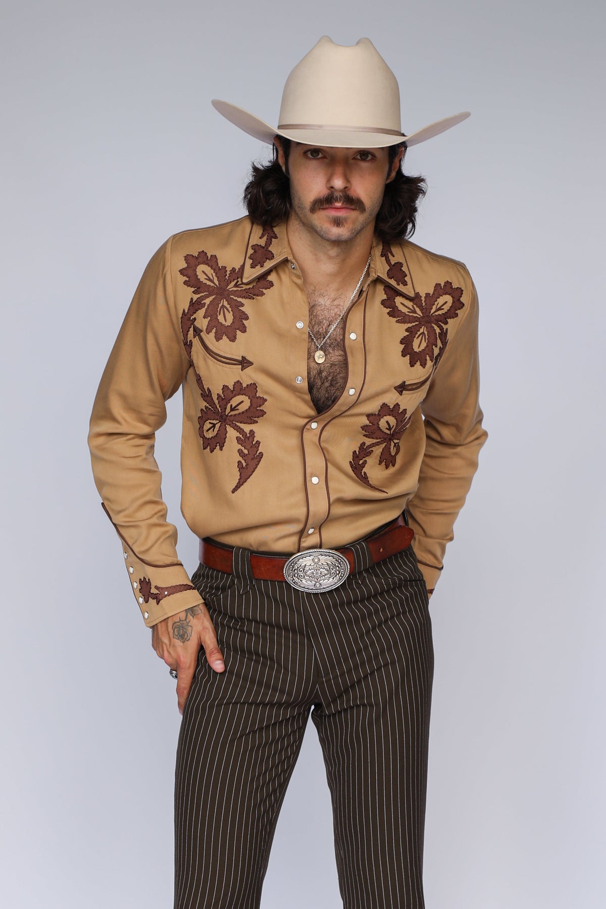 Man in a cream cowboy hat wearing the H Bar C Terlingua shirt in tan with brown scroll embroidery, an H Bar C archive remake, with pinstripe pants.