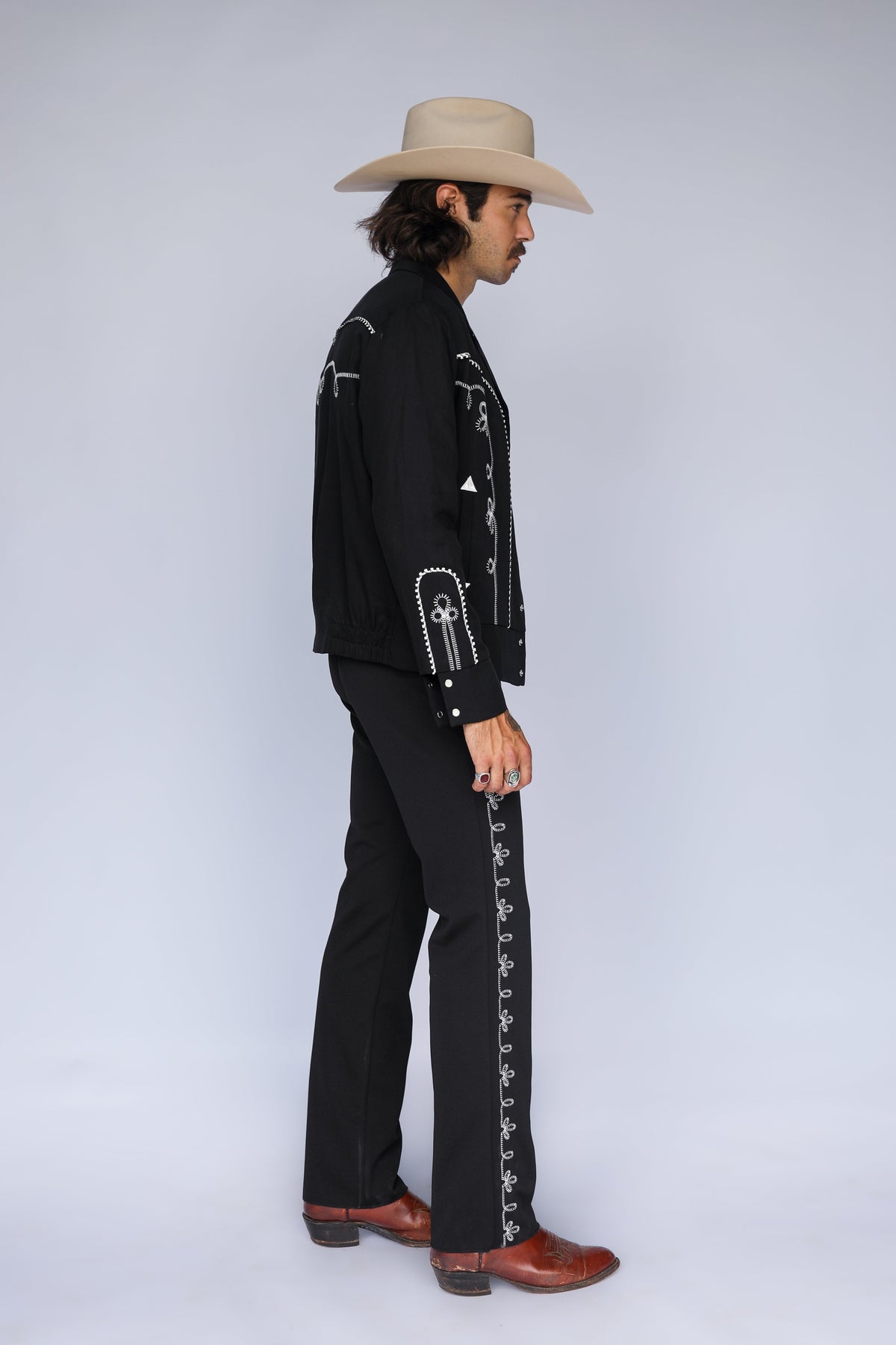 Profile of a man wearing the H Bar C Bolero pants and Bolero jacket in black with white embroidery and a cream cowboy hat.