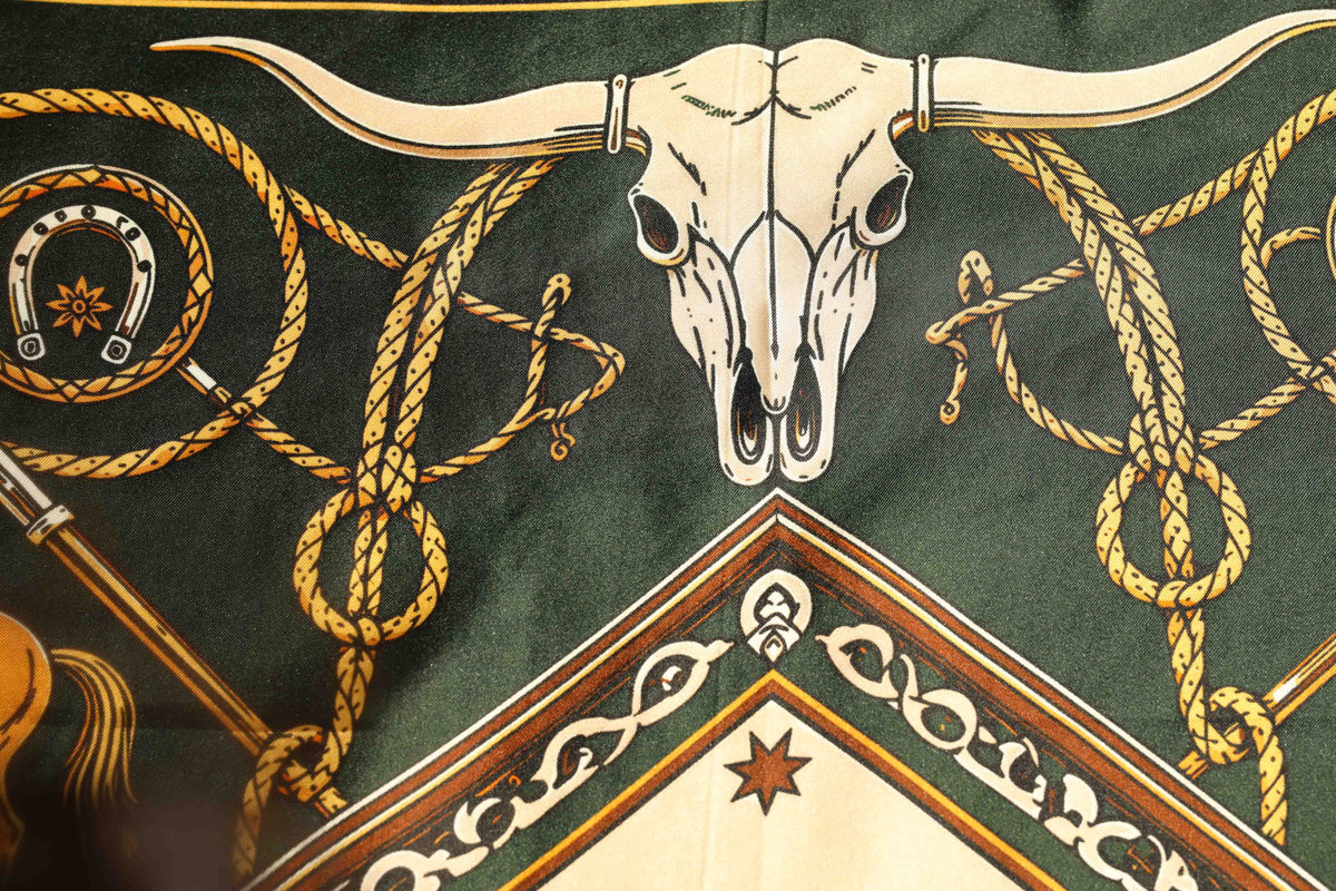 Close-up of the H Bar C Green silk scarf showing a longhorn steer skull, gold lariat rope, and horseshoe motifs on hunter green silk.
