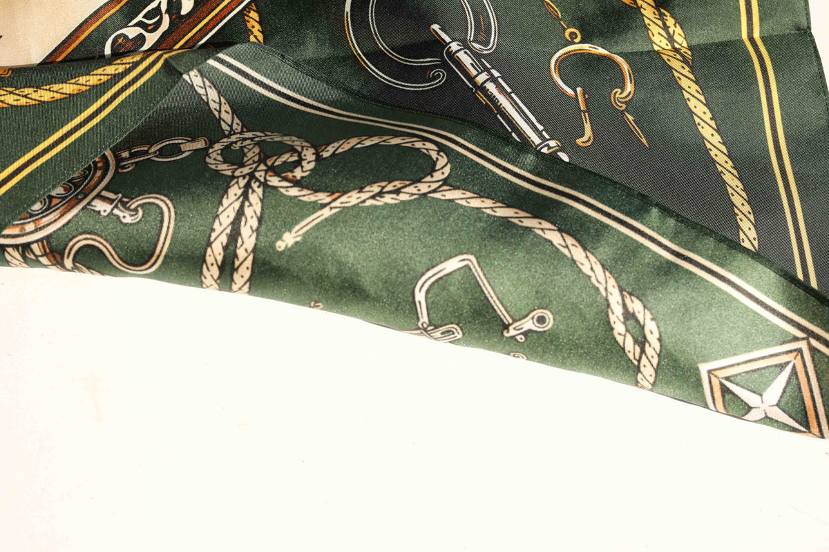 Draped edge of the H Bar C Green silk scarf showing the hand-finished hem, gold border stripe, rope, and horse bit details.