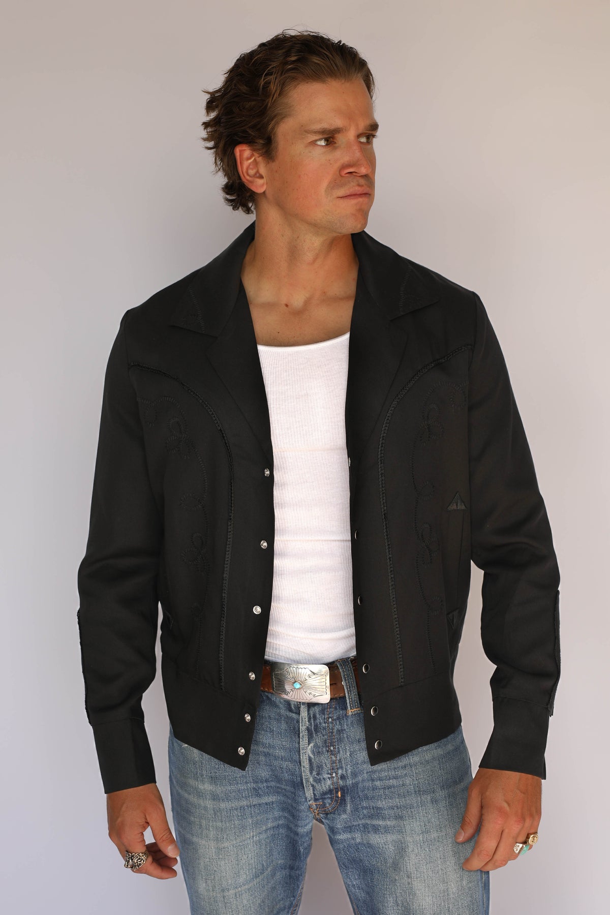 Man wearing the H Bar C Bolero men's jacket in black on black with tonal chain stitch embroidery, over a white tank with jeans.