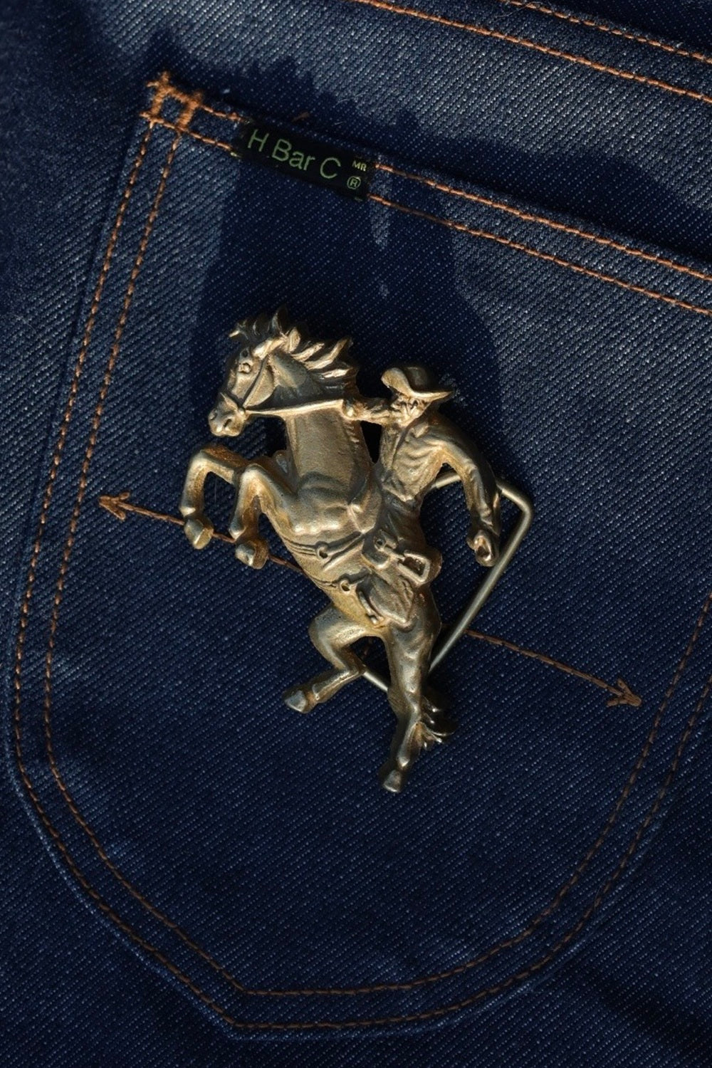 Detail of the H Bar C gold cowboy belt buckle with bucking bronco on raw denim.