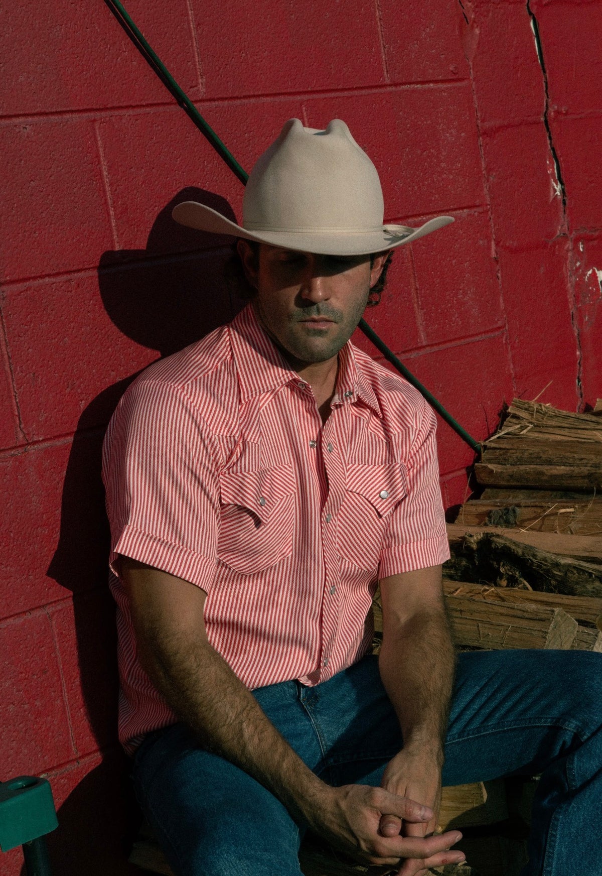 Man sitting in front of a red wall wearing the H Bar C Newport red stripe western shirt and cowboy hat.