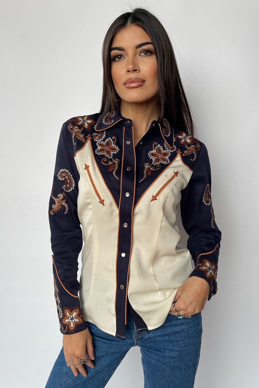 Woman wearing the H Bar C Aspen women's shirt in off white with navy yoke and sleeves, copper floral embroidery, and arrow pockets, designed by Vera Vasiley.