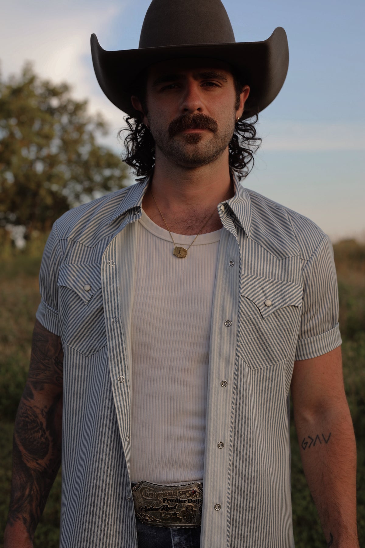 H Bar C Newport gray striped short sleeve snap shirt worn open over a white tee with a black cowboy hat outdoors at sunset.