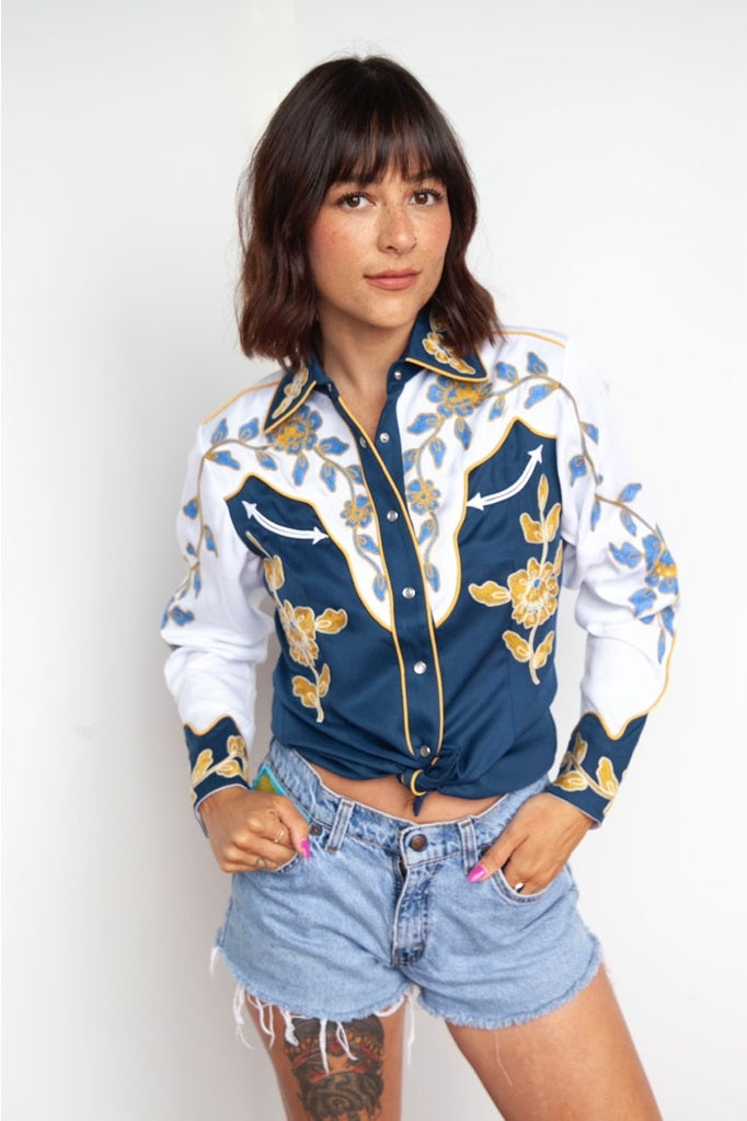 Woman wearing the H Bar C Charleston women's shirt in blue with white yokes and gold chain stitch flowers, with denim shorts.