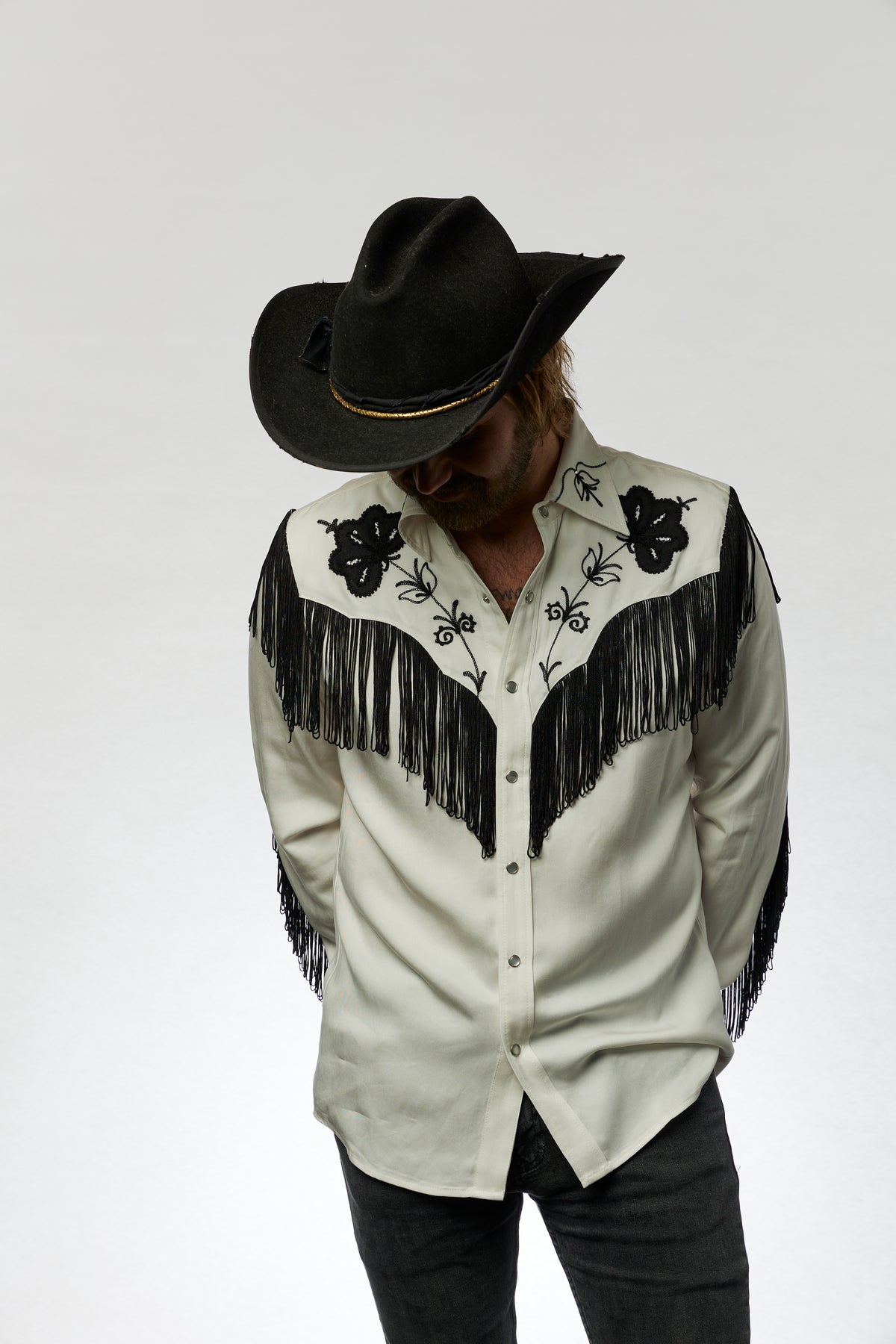 Man in a black cowboy hat looking down in the H Bar C Taos white and black fringe shirt.